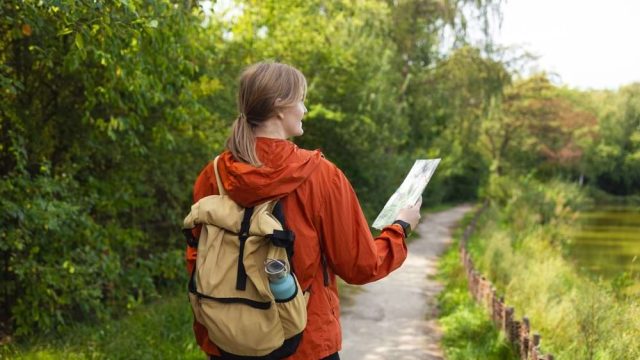 A woman with a rucksack reading a map on a path beside the water