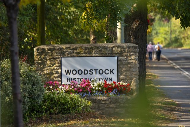 The Woodstock Historic Town sign on a stone wall with flower beds, beside a tree-lined road