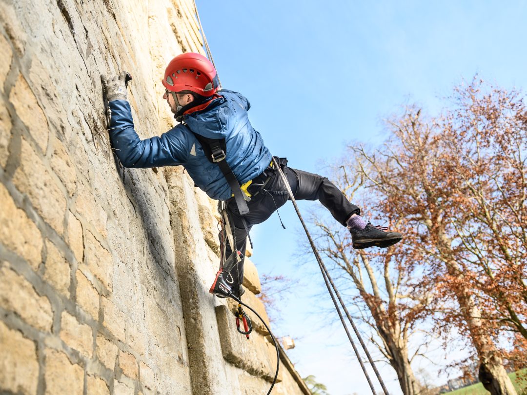 A climber in a red helmet and blue jacket abseiling down the stonework of the Grand Bridge