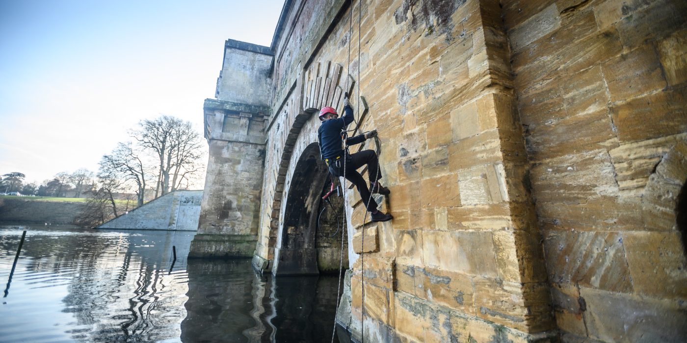 An ecologist abseiling down the side of the Grand Bridge beside an arch, with the lake below