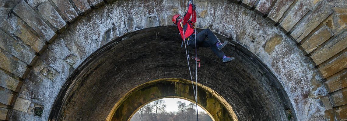 An ecologist in a red helmet abseiling inside an arch of the Grand Bridge, above the water