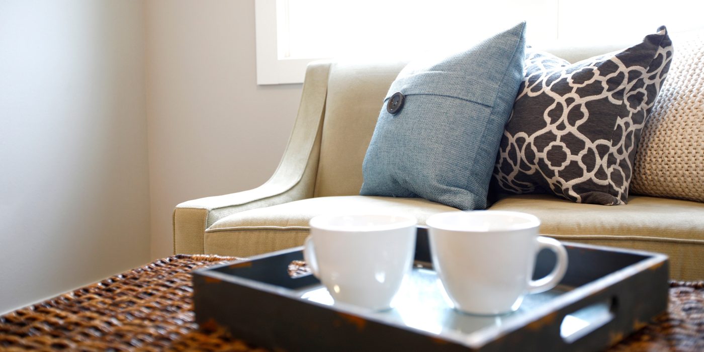 Two white mugs on a tray on a wicker table in front of a sofa with cushions
