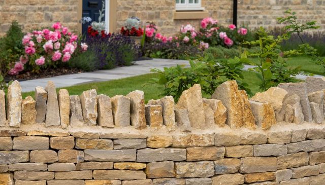 A new dry stone wall in front of a planted garden with pink peonies and a stone house