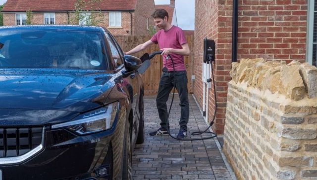 A man charging an electric car from a wall charger on the side of a new brick home
