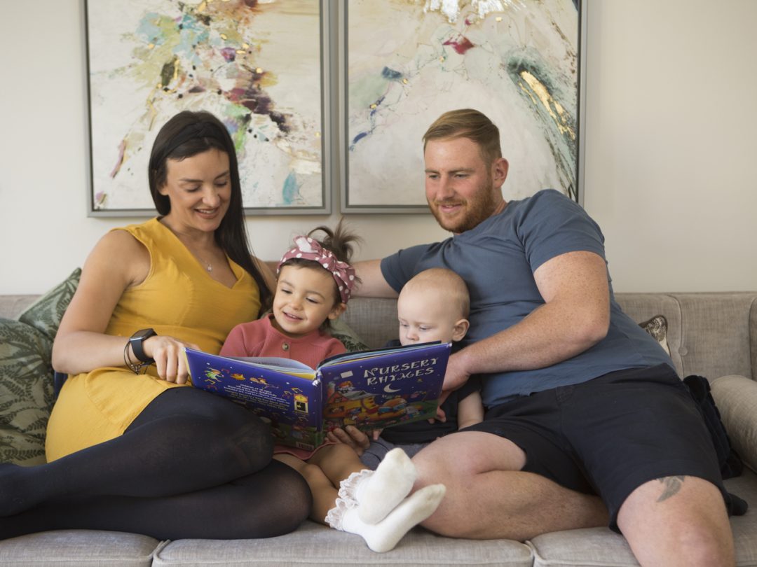 Parents reading a picture book with their young daughter and baby on a sofa