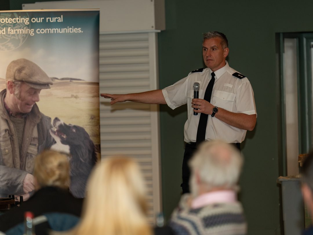 A police officer with a microphone speaking to the farming conference audience beside a rural crime banner