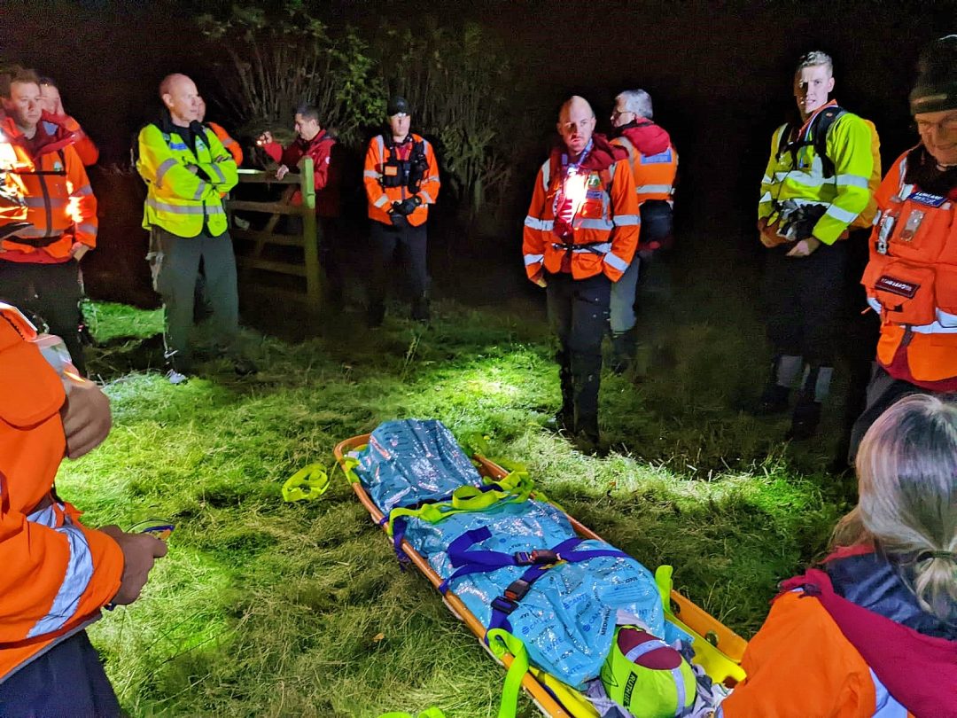 Lowland Rescue volunteers around a casualty strapped to a stretcher on grass at night, lit by head torches