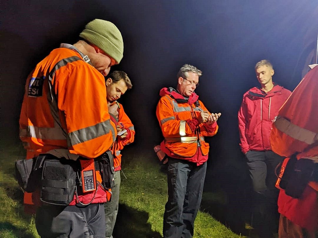 Lowland Rescue search team members in high-visibility jackets gathered on grass at night for a briefing