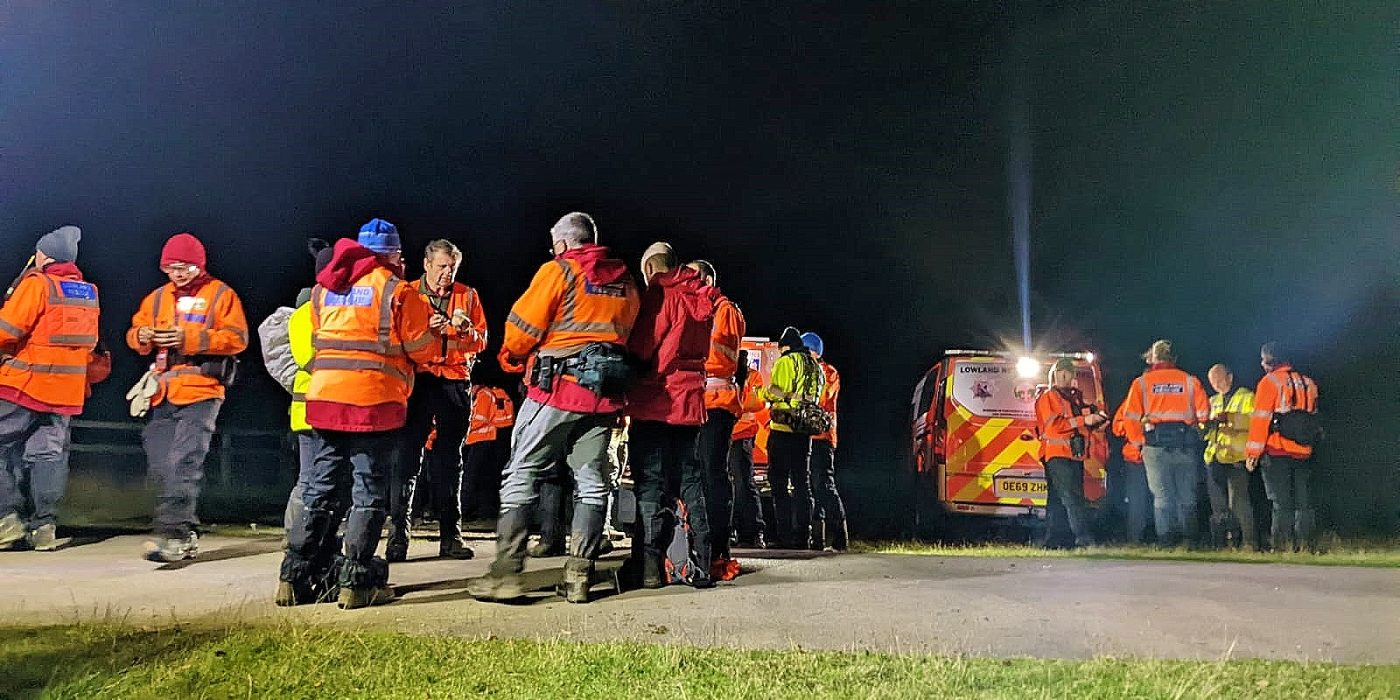 Lowland Rescue search teams assembled on a track at night beside a rescue vehicle under a floodlight