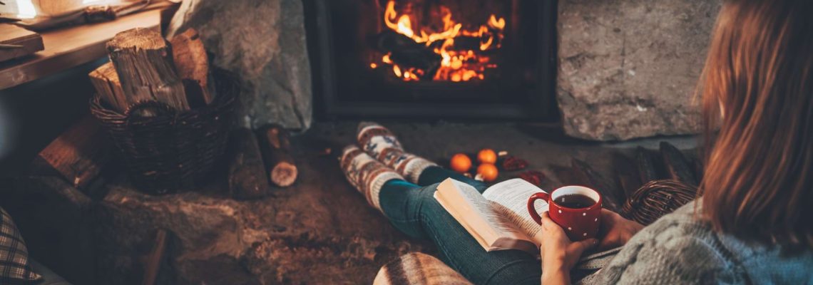 A woman reading a book with a red mug, her feet up in front of a log fire
