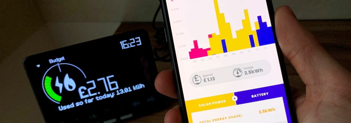 A hand holding a phone showing an energy usage app next to a smart meter display