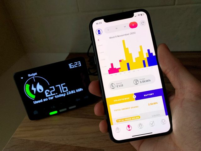 A hand holding a phone showing an energy usage app next to a smart meter display