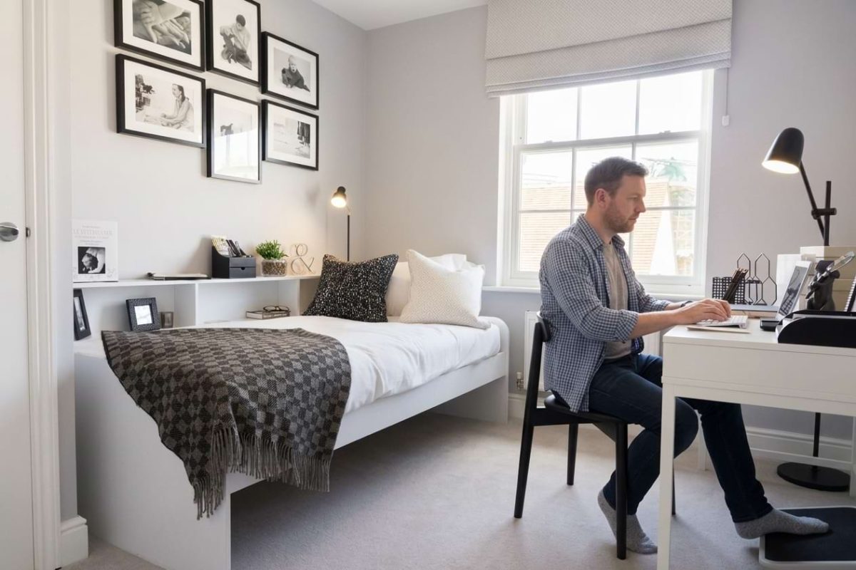 A man working at a laptop in a small study bedroom with a single bed and framed photos