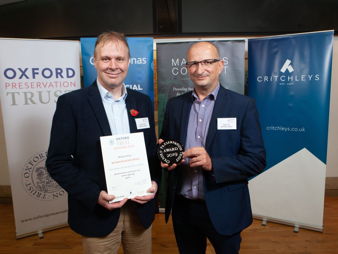 Two men holding an Oxford Preservation Trust Award certificate and plaque in front of sponsor banners