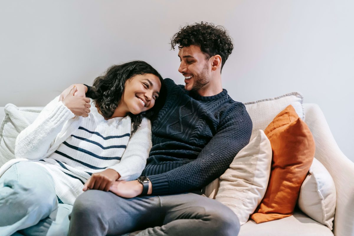 A smiling couple relaxing together on a white sofa with orange and cream cushions