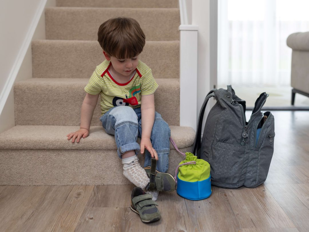 A small boy sitting on the stairs putting on his shoes, next to a rucksack and lunchbox