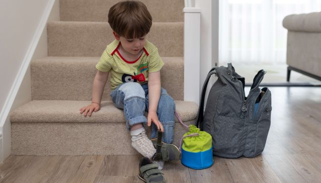 A small boy sitting on the stairs putting on his shoes, next to a rucksack and lunchbox