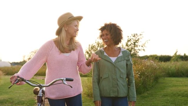 Two women laughing together in a meadow, one pushing a bicycle