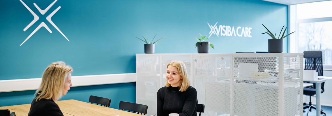 Two Visiba Care staff talking at a table in their bright office, with the company logo on a teal wall