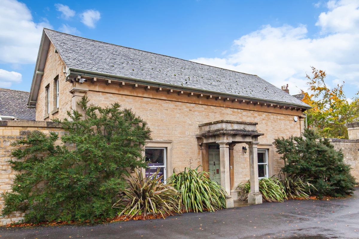 Woodstock Lodge, a single-storey stone building with a columned porch, behind a stone wall and shrubs