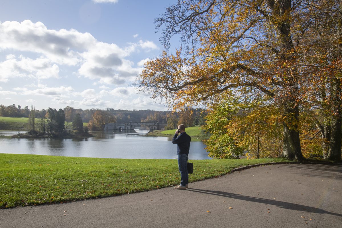 A man on the phone on a path above the lake in Blenheim Park, with autumn trees and the Grand Bridge in the distance