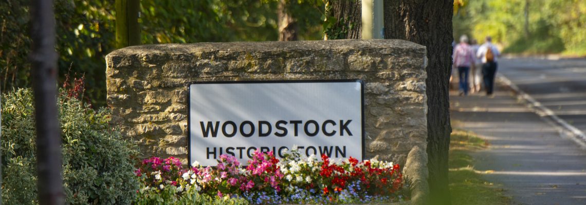 The Woodstock Historic Town sign on a stone wall with flower beds, beside a tree-lined road