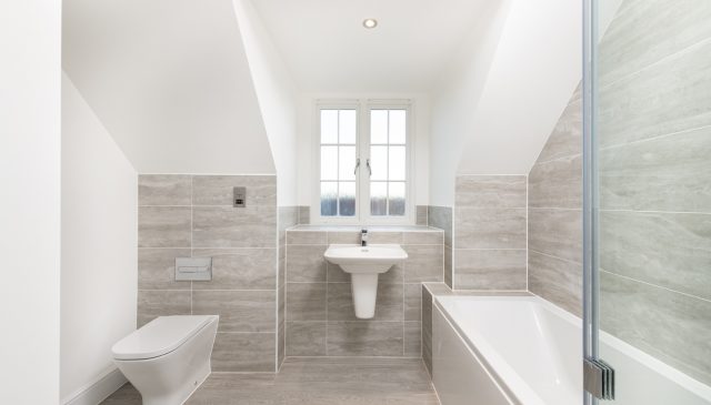 A bathroom tiled by Minoli, with a wall-hung toilet and basin, a bath and a window under the eaves