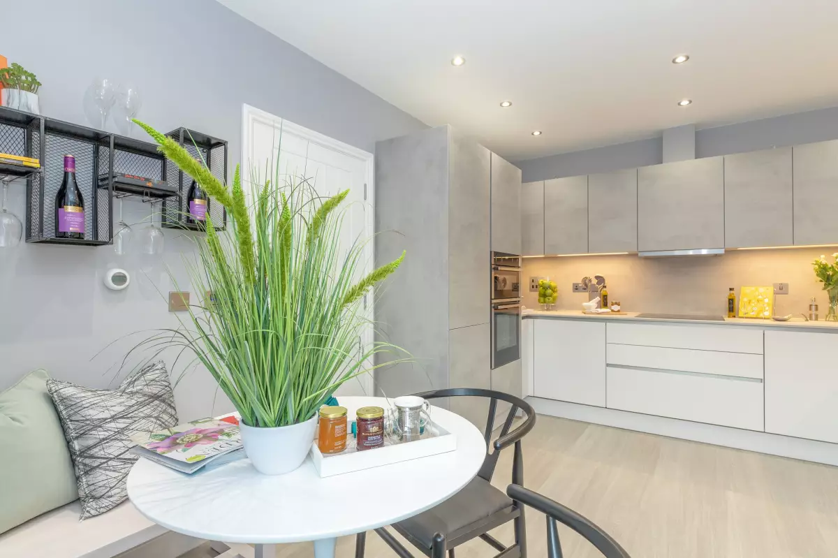 A Minoli-fitted kitchen with white units, a round breakfast table and wine bottles on wall shelves